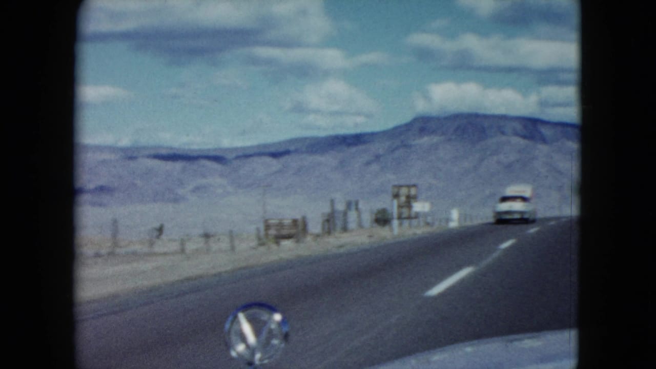 1959 Nevada road footage shows a white car ahead on the highway, with roadside signs and mountains in the distance.