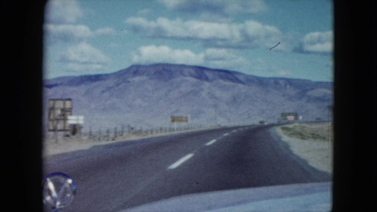Open desert road curves ahead with mountains, clouds, and roadside signs visible.