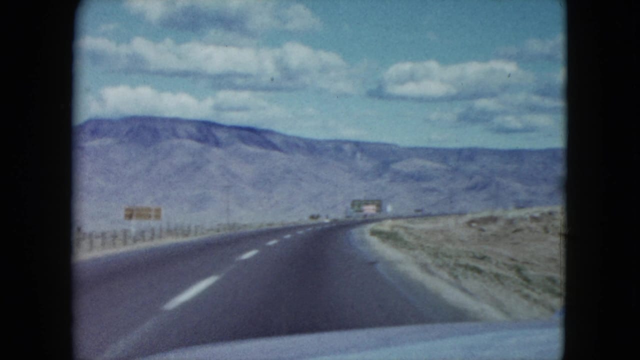 Forward-facing 8mm view of a Nevada desert road bending slightly, with mountains and sparse signs ahead.