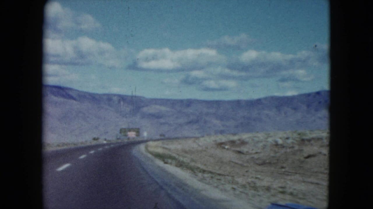 The 1959 Nevada road scene continues from a car, showing an empty highway, desert shoulder, and mountain backdrop.