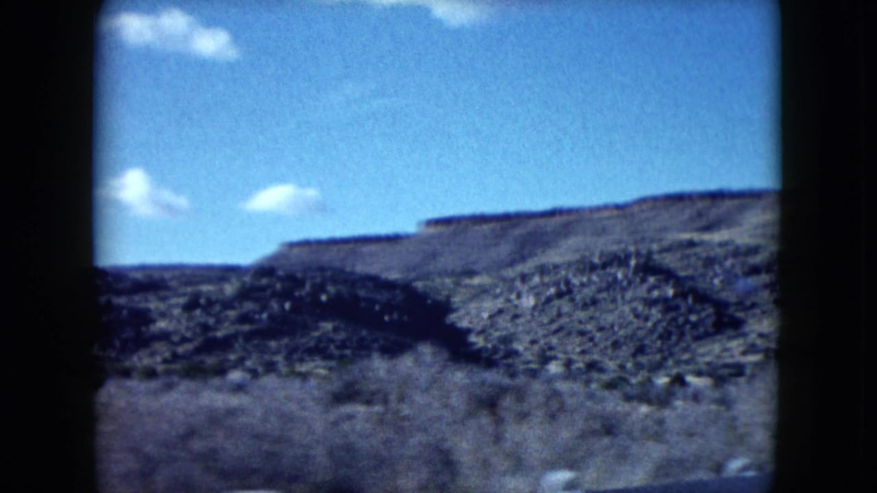 Rocky Nevada desert terrain passes under a bright blue sky, framed by the dark edge of the moving vehicle or film gate.