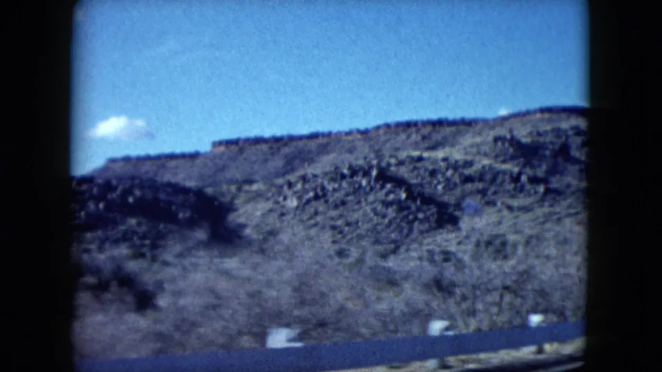 From a moving car, blurred roadside brush crosses the foreground with rocky Nevada hills and blue sky beyond.