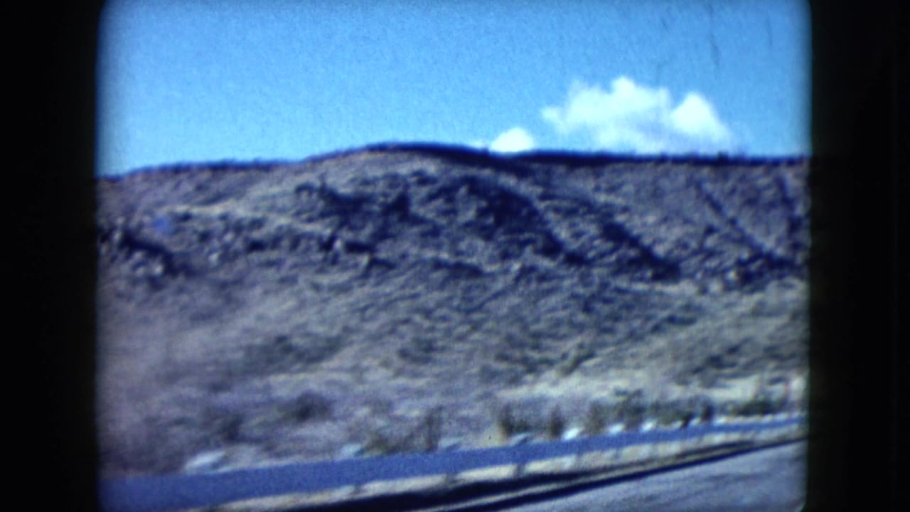 1959 Nevada 8mm frame showing a rocky desert hill, roadside guardrail, and blue sky from a moving vehicle.