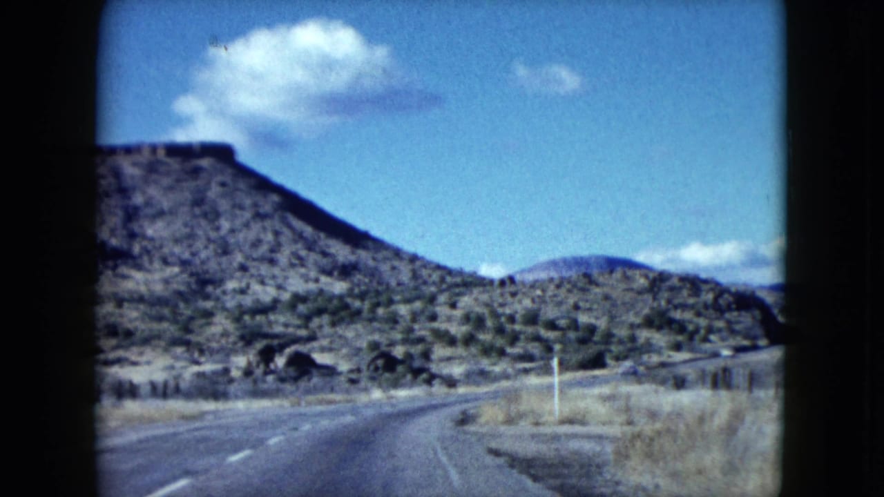 1959 Nevada 8mm frame of a curving desert highway bordered by dry grass, low shrubs, and rocky hills beneath a blue sky.