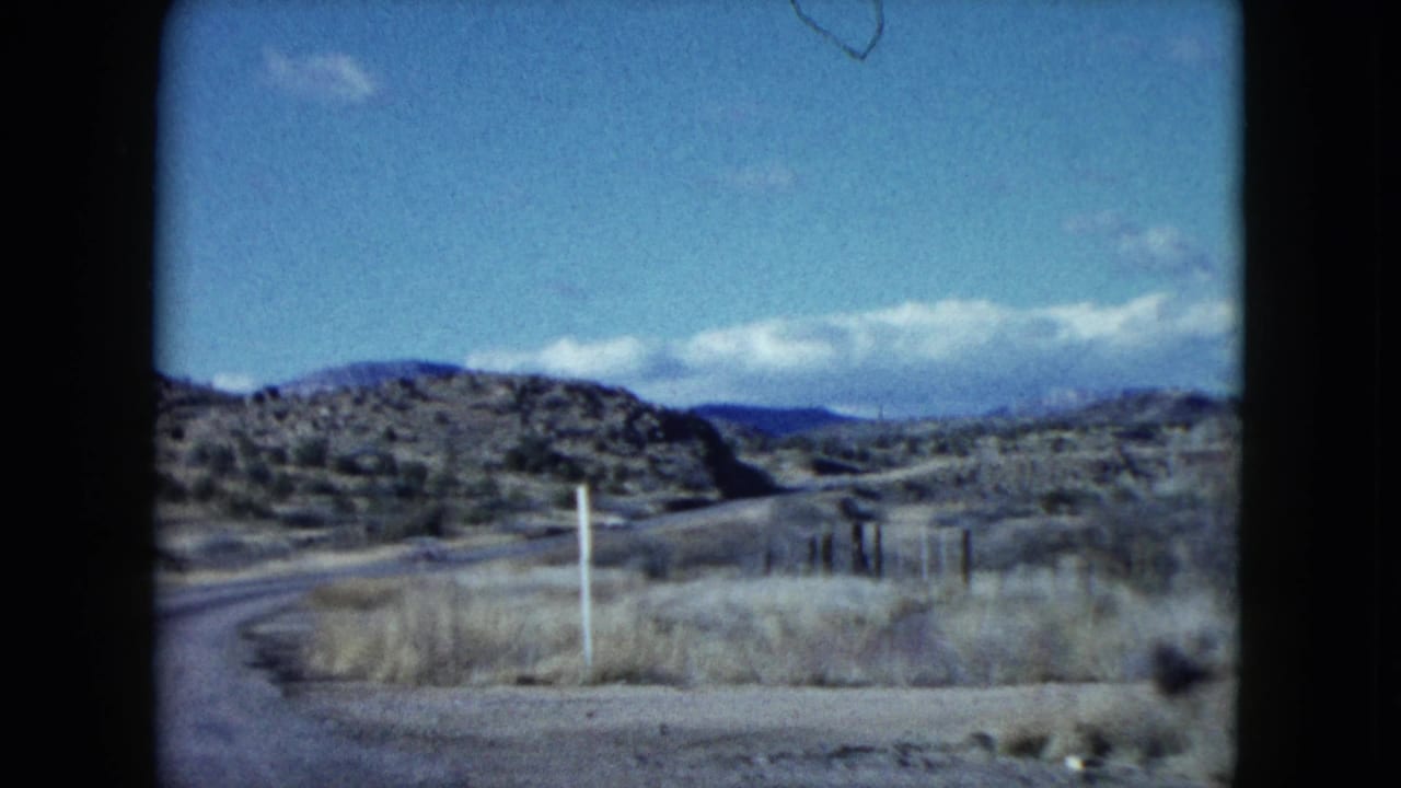 1959 Nevada roadside view showing dry grass, fence or guard posts, low desert hills, and distant clouds from a moving car.
