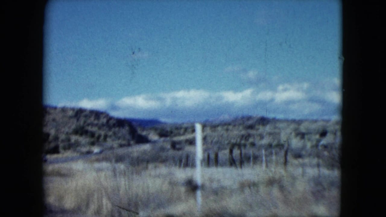 1959 Nevada home-movie frame from a moving car, showing arid roadside land, fence posts, low mountains, and a clouded blue sky.