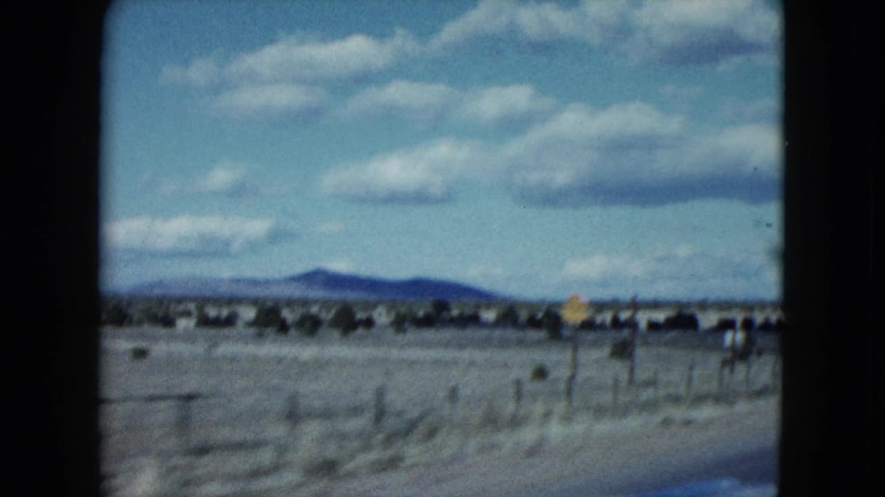 Desert landscape seen through car-window framing with low mountains, fence line, and roadside sign.