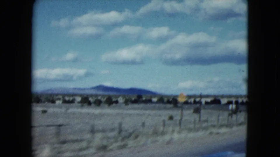 1959 Nevada driving view with black car-window borders, open desert land, low mountains, fence line, and a small sign in the distance.