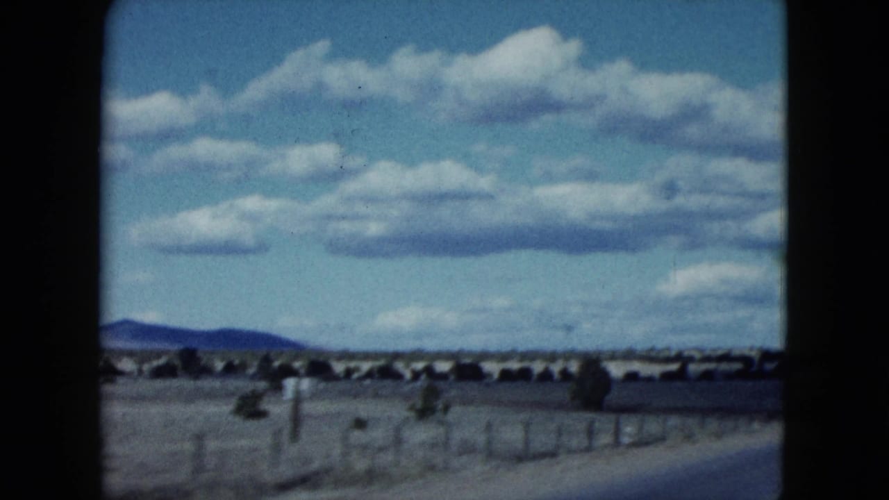 1959 Nevada roadside frame showing dry open land, fence posts, low hills, and broad clouds from a moving vehicle.