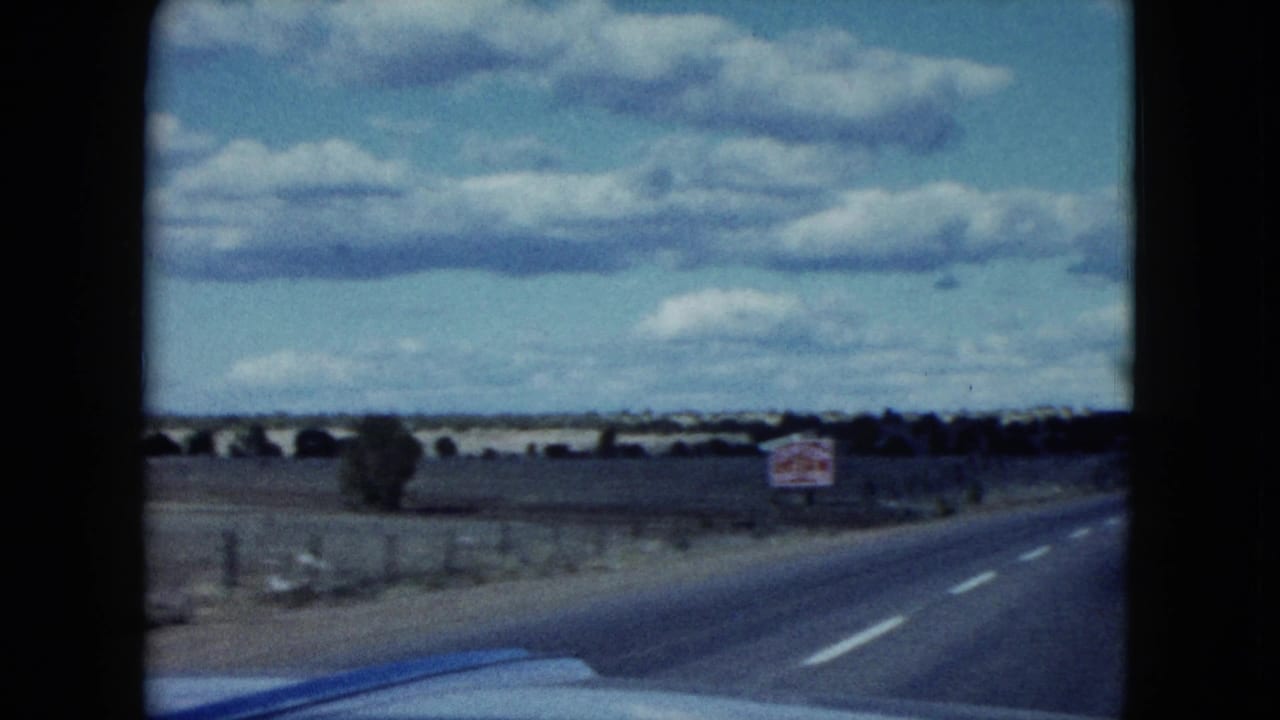 1959 Nevada home-movie frame of a two-lane blacktop road curving through desert countryside with a sign, fence, and car hood visible.