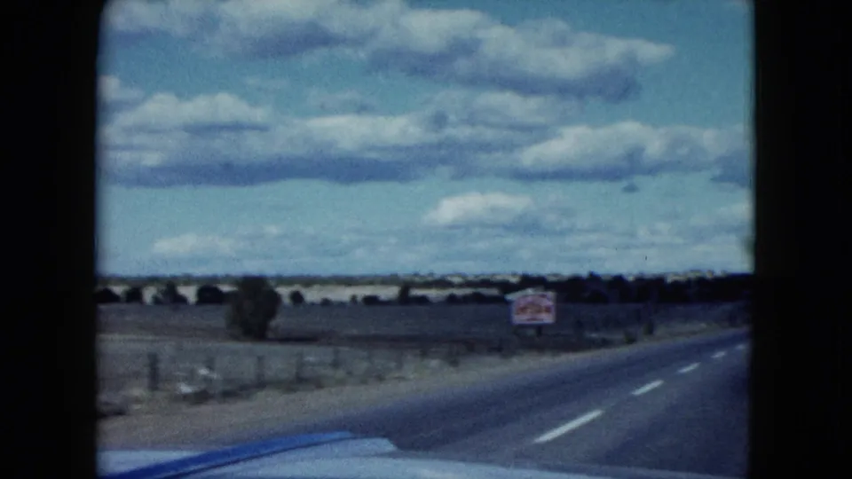 1959 Nevada home-movie frame of a two-lane blacktop road curving through desert countryside with a sign, fence, and car hood visible.