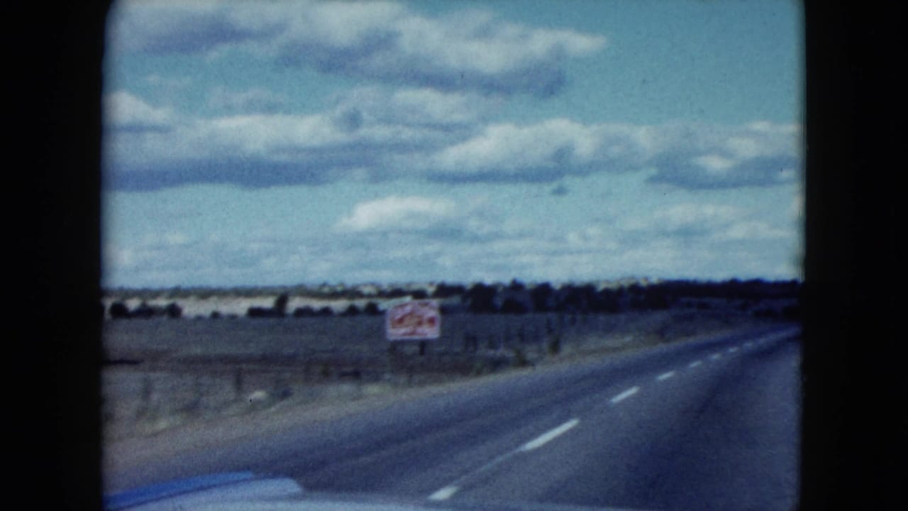 1959 Nevada 8mm view from a car on a quiet paved road bordered by scrubland, fencing, and a distant roadside sign.