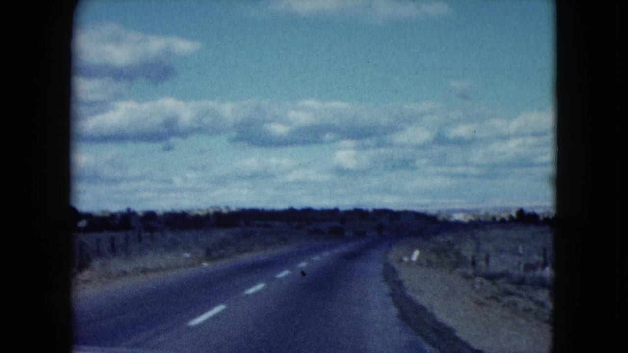 Curving empty road bordered by scrub and fence posts beneath a blue sky with clouds.