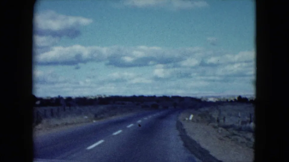 1959 Nevada home-movie frame showing an empty road curving through dry open country with fence lines and scattered clouds.