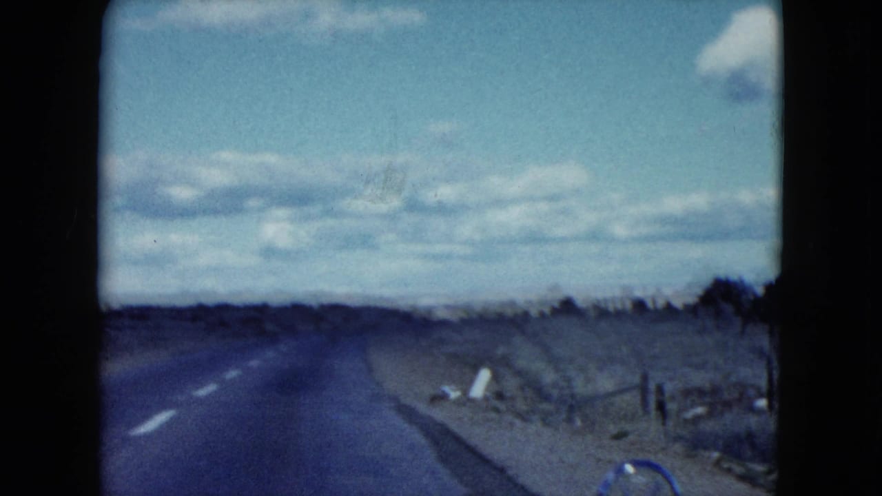 1959 Nevada 8mm frame from a moving car, looking down a quiet road with scrub, fencing, and soft home-movie exposure.