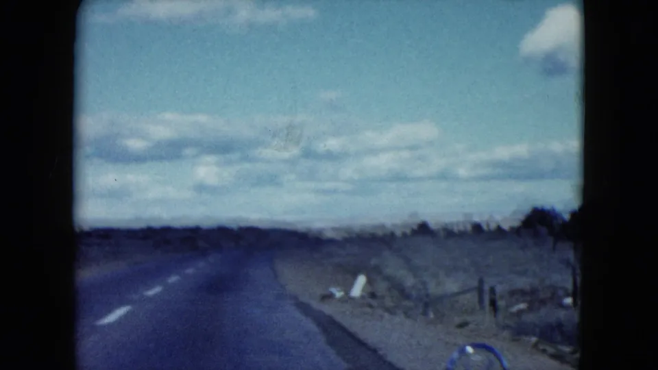 1959 Nevada 8mm frame from a moving car, looking down a quiet road with scrub, fencing, and soft home-movie exposure.