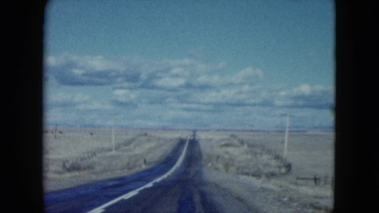 1959 Nevada road-trip frame showing a long empty road stretching across flat arid land with utility poles and distant horizon.
