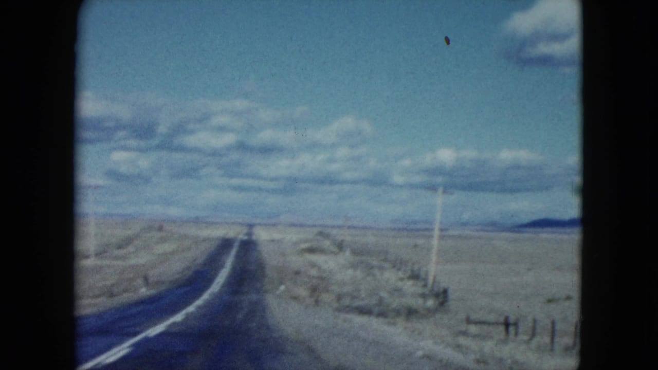 1959 Nevada 8mm frame showing a two-lane desert road, roadside fence, utility poles, scrubland, and mountains on the horizon.