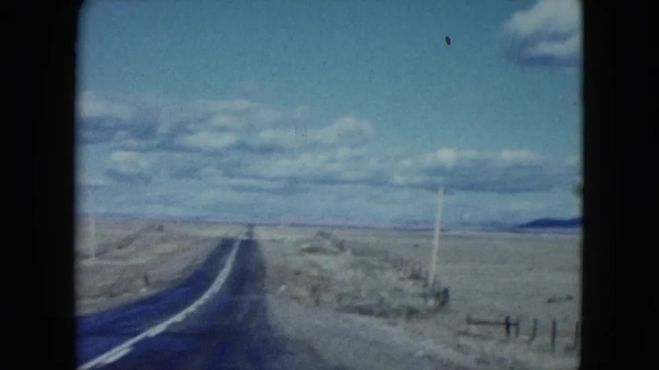 1959 Nevada 8mm frame showing a two-lane desert road, roadside fence, utility poles, scrubland, and mountains on the horizon.