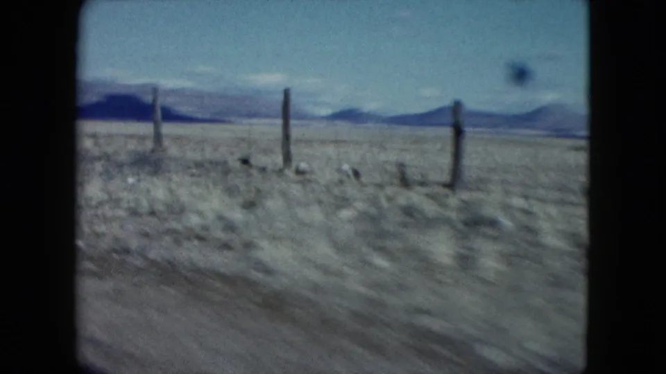 1959 Nevada 8mm frame showing a blurred desert roadside with wooden fence posts, low mountains, and open blue sky.