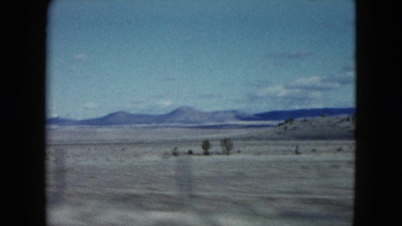 Open desert plain with distant mountains, low scrub, and pale blue sky.