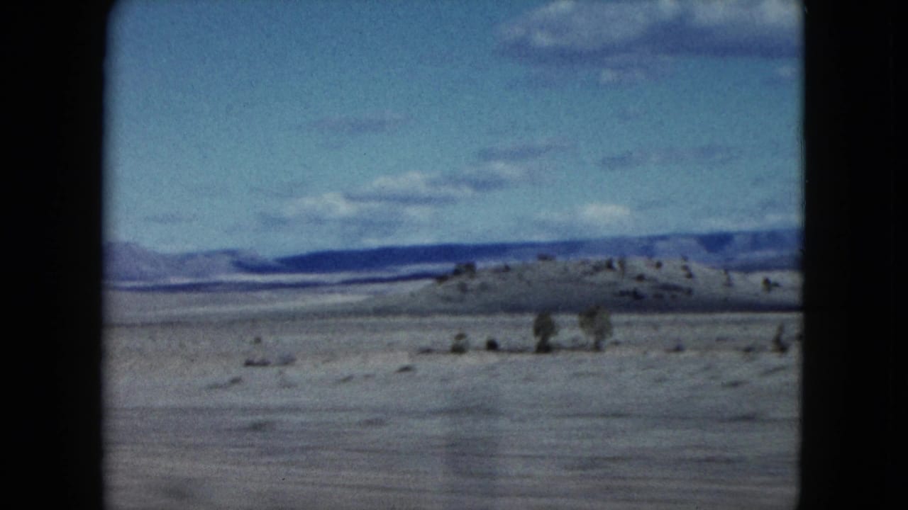 1959 Nevada 8mm frame of a desert basin with scattered shrubs, a low ridge, distant mountains, and soft clouds.