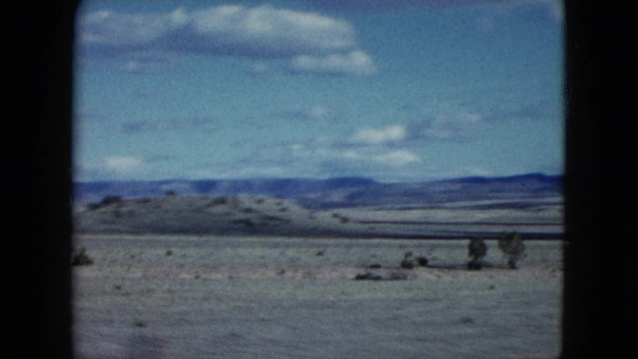 1959 Nevada landscape frame showing sparse desert brush, a low hill, distant ridgeline, and bright blue sky with clouds.