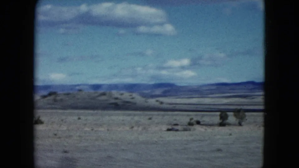1959 Nevada landscape frame showing sparse desert brush, a low hill, distant ridgeline, and bright blue sky with clouds.