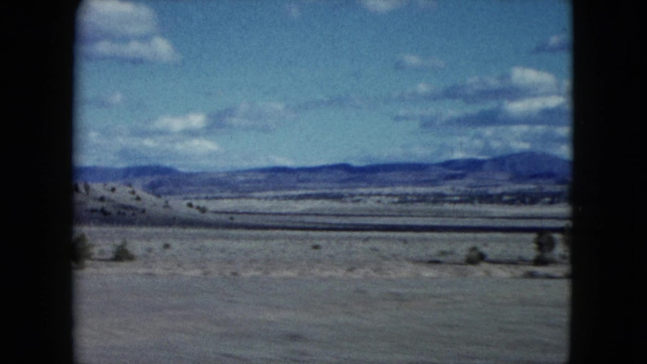 Open desert plain with scattered shrubs, distant low mountains, blue sky, and film gate edges.