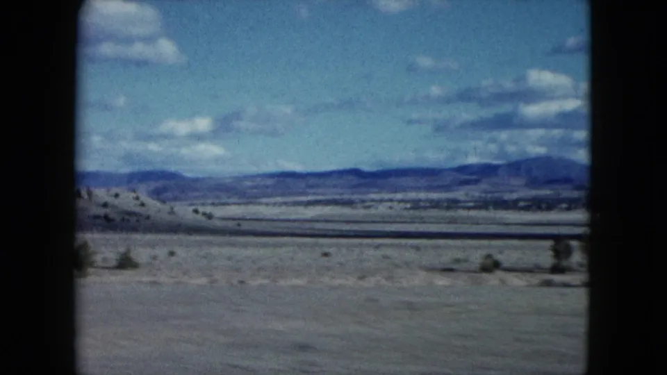 1959 Nevada 8mm frame showing an arid plain, scattered desert vegetation, low hills, distant mountains, and a bright clouded sky.