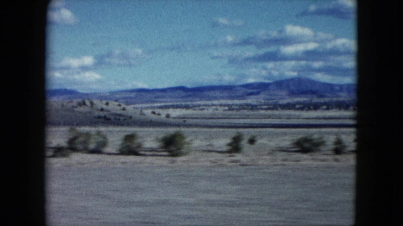 1959 Nevada 8mm frame with a broad desert foreground, blurred shrubs, layered low hills, distant mountains, and bright clouds overhead.