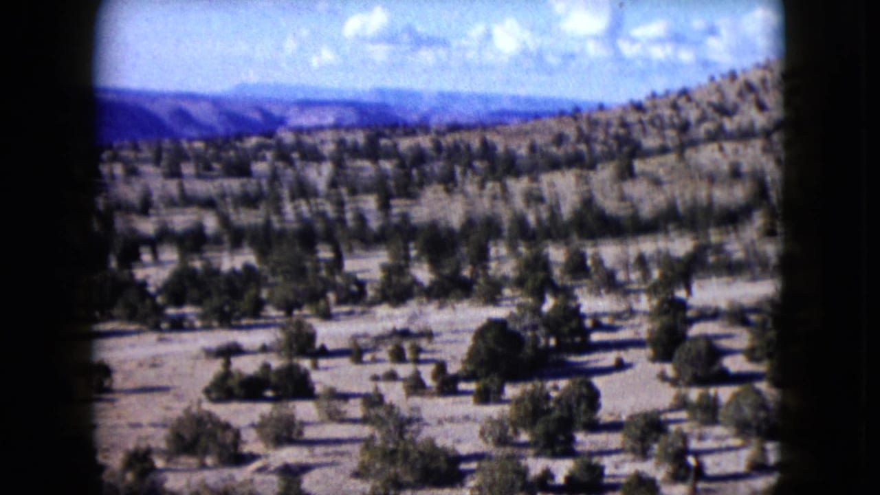 1959 Nevada 8mm frame showing a dry sloping landscape with scattered vegetation, open valley ground, distant ridges, and a bright clouded sky.