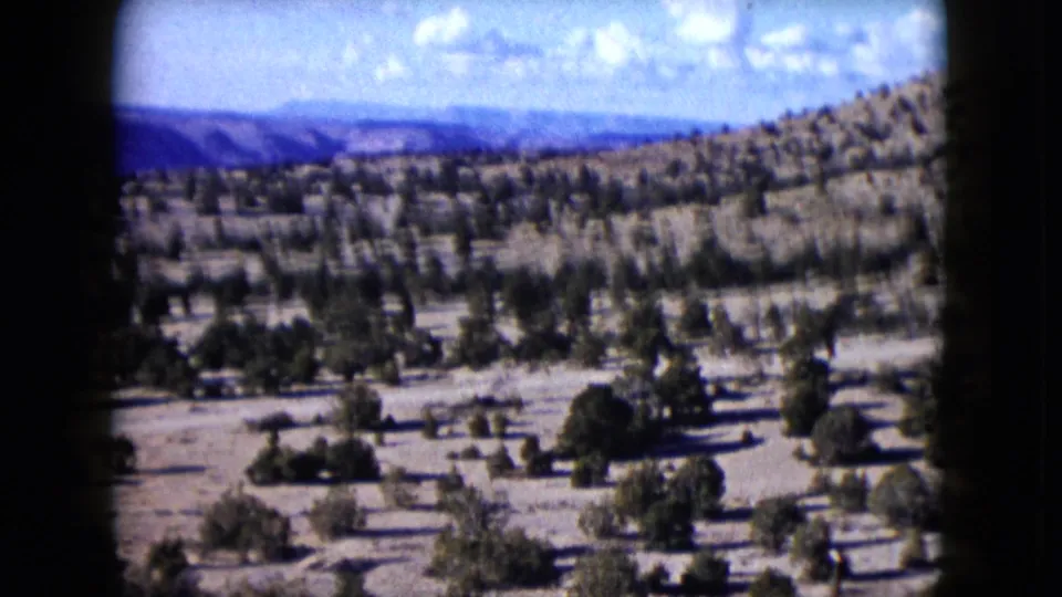 1959 Nevada 8mm frame showing a dry sloping landscape with scattered vegetation, open valley ground, distant ridges, and a bright clouded sky.