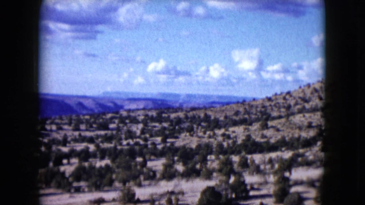 1959 Nevada 8mm frame looking across a dry valley and shrub-covered hillside toward distant blue ridges beneath scattered clouds.