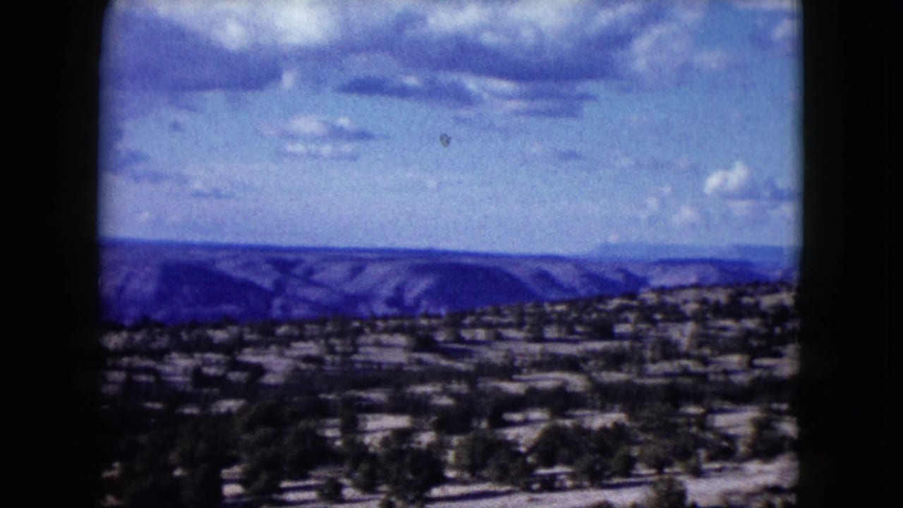 1959 Nevada 8mm frame with a broad arid foreground, scattered shrubs, distant mesas, and soft clouds in blue sky.
