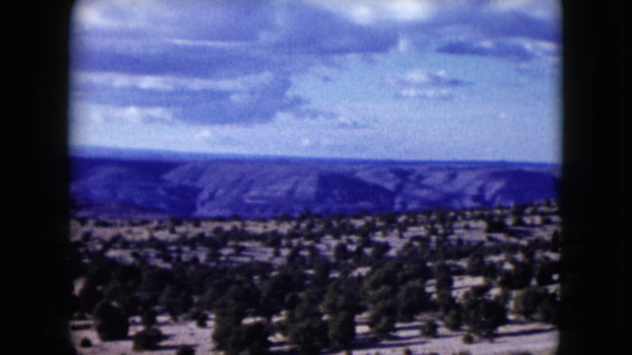 1959 Nevada landscape frame showing hazy mesas beyond scrub-covered desert ground beneath layered clouds.