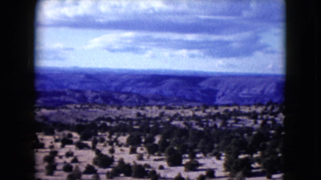 1959 Nevada home-movie still of open desert vegetation leading to blue-toned mesas under a bright cloudy sky.
