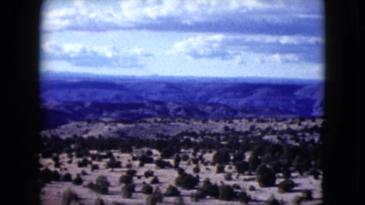 1959 Nevada home-movie frame showing dry scrubland, scattered trees, and distant blue ridges beneath a bright cloudy sky.