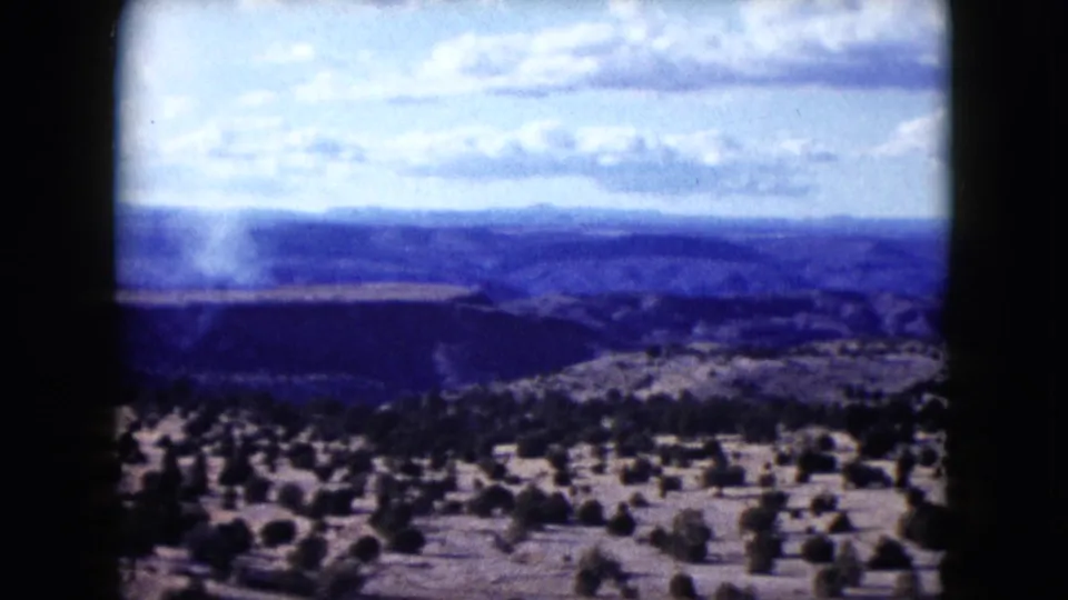 1959 Nevada landscape view with scrub trees, distant mountain ridges, and a faint pale smoke plume near the valley.
