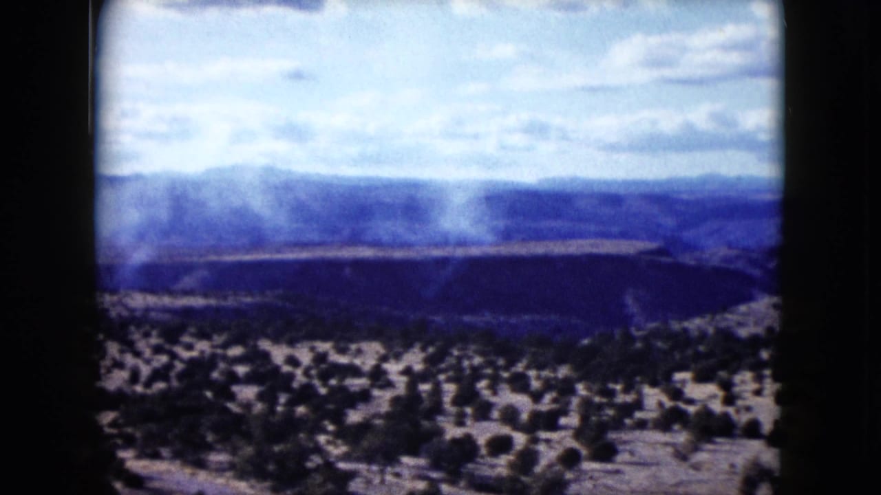 1959 Nevada home-movie frame showing a white smoke plume rising across an arid valley below distant blue hills.