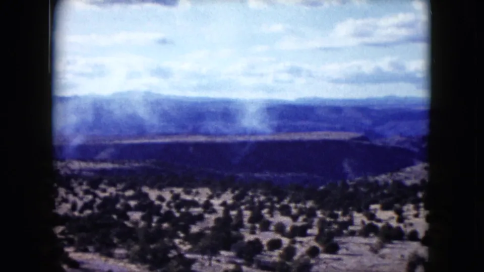 1959 Nevada home-movie frame showing a white smoke plume rising across an arid valley below distant blue hills.