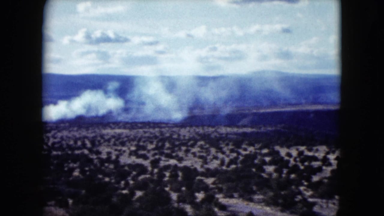 1959 Nevada landscape frame with a larger white smoke plume over scrubland, backed by distant hills and a bright sky.