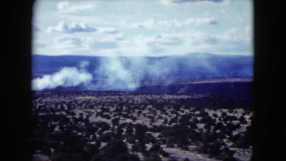 1959 Nevada landscape frame with a larger white smoke plume over scrubland, backed by distant hills and a bright sky.