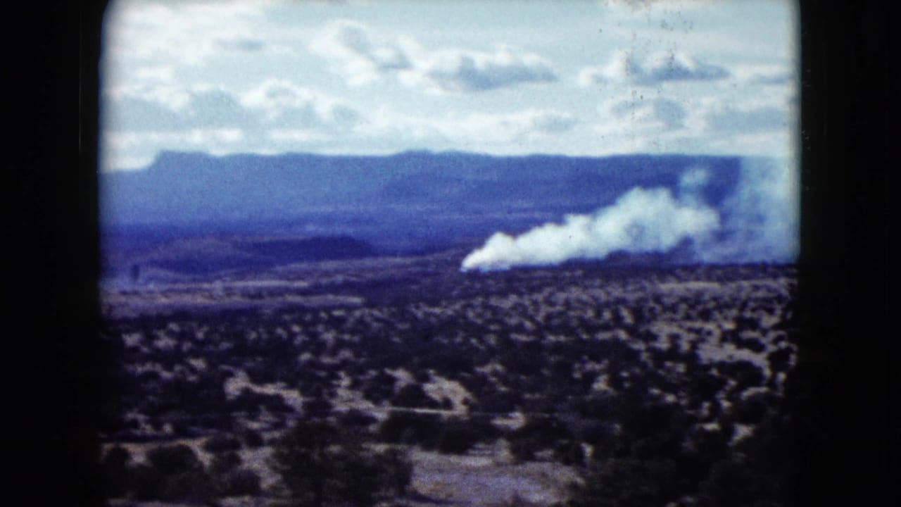 White smoke plume spreads across a scrub desert with mountains beyond.