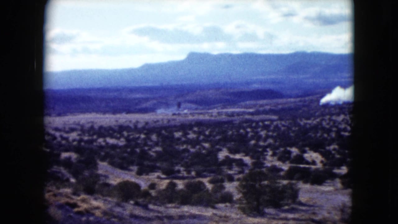 1959 Nevada 8mm frame showing scrubland, distant mountains, and a partial white smoke plume at the edge of the view.