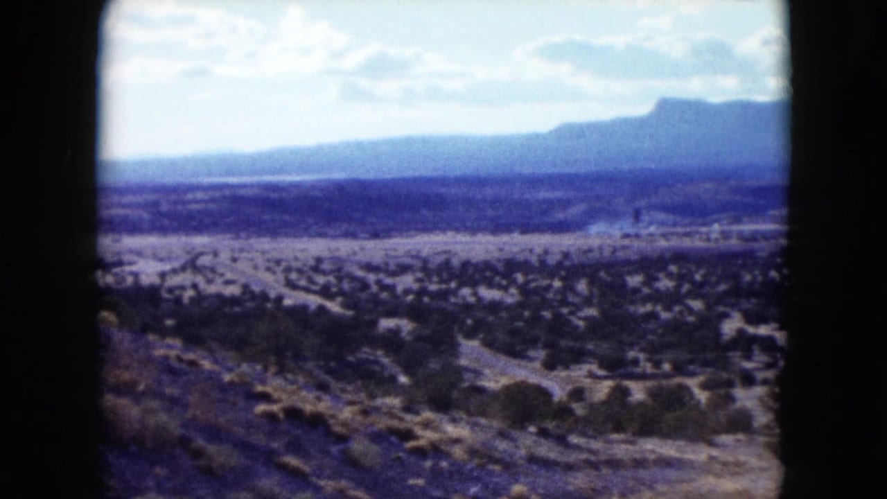 1959 Nevada frame showing a wide arid landscape, dark shrubs, distant mountains, and faint smoke far across the valley.