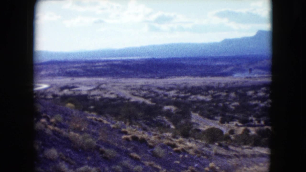 1959 Nevada home-movie frame showing an arid valley, scrub vegetation, low hills, and distant mountains from a roadside viewpoint.