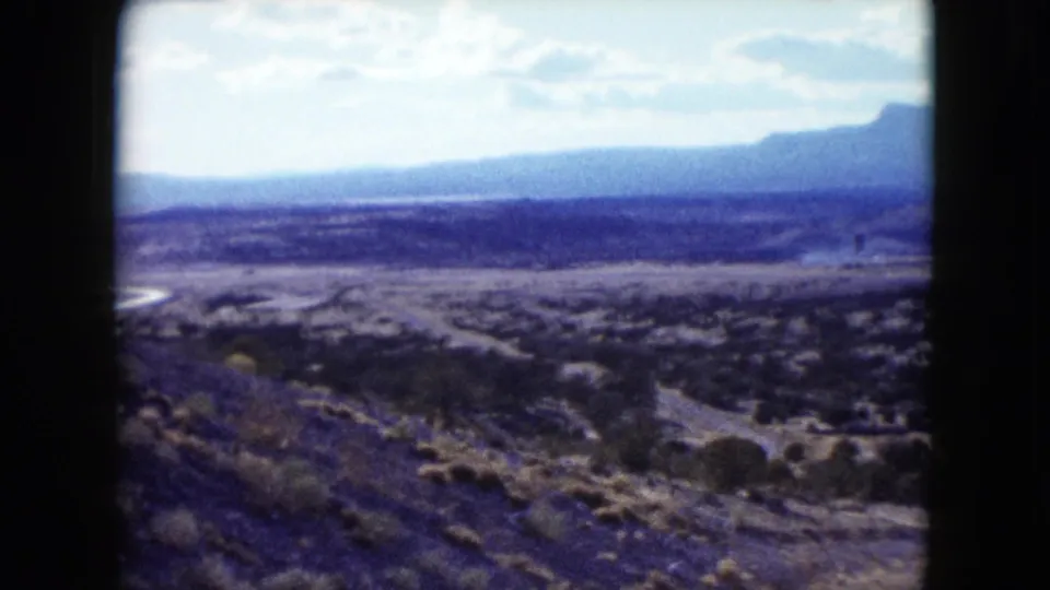 1959 Nevada home-movie frame showing an arid valley, scrub vegetation, low hills, and distant mountains from a roadside viewpoint.