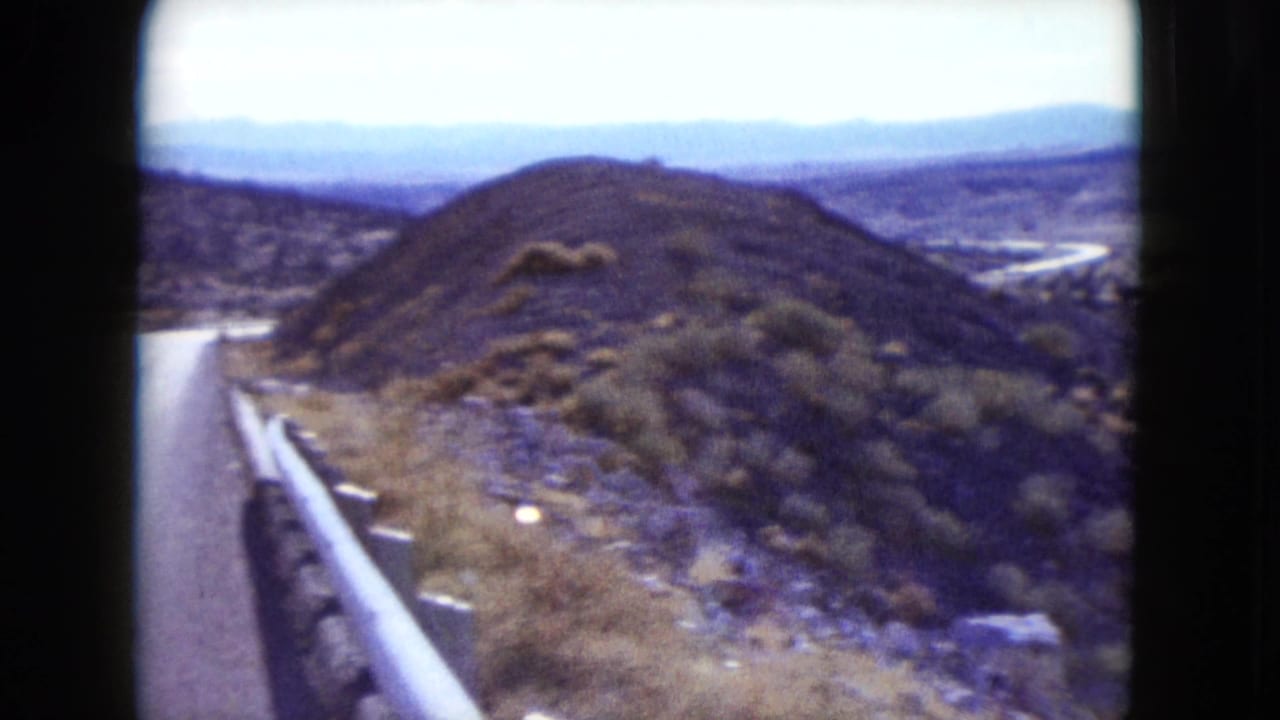 1959 Nevada frame from a moving vehicle, showing a roadside guardrail, dry brush-covered hills, and distant arid terrain.