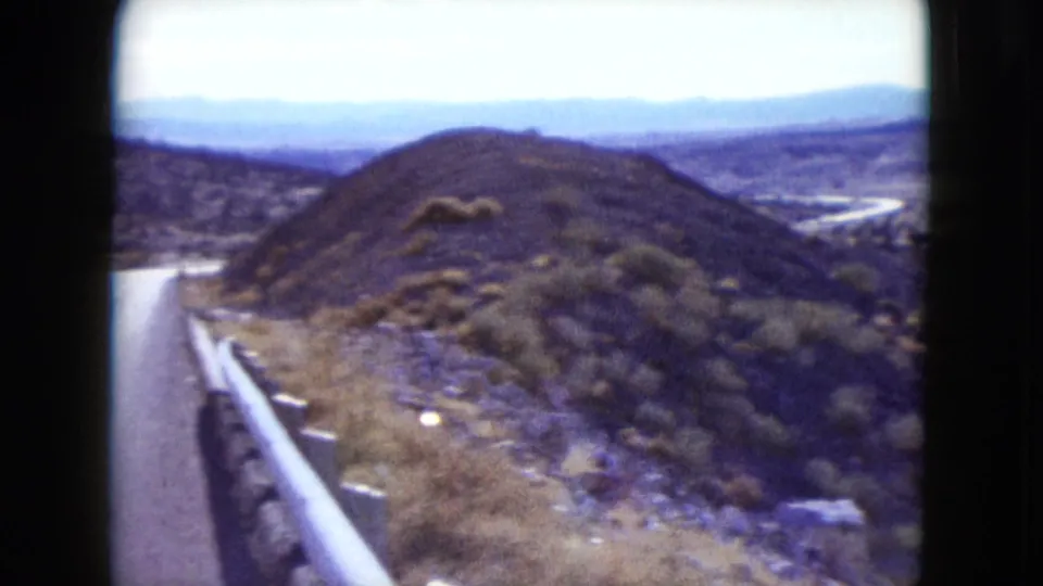 1959 Nevada frame from a moving vehicle, showing a roadside guardrail, dry brush-covered hills, and distant arid terrain.